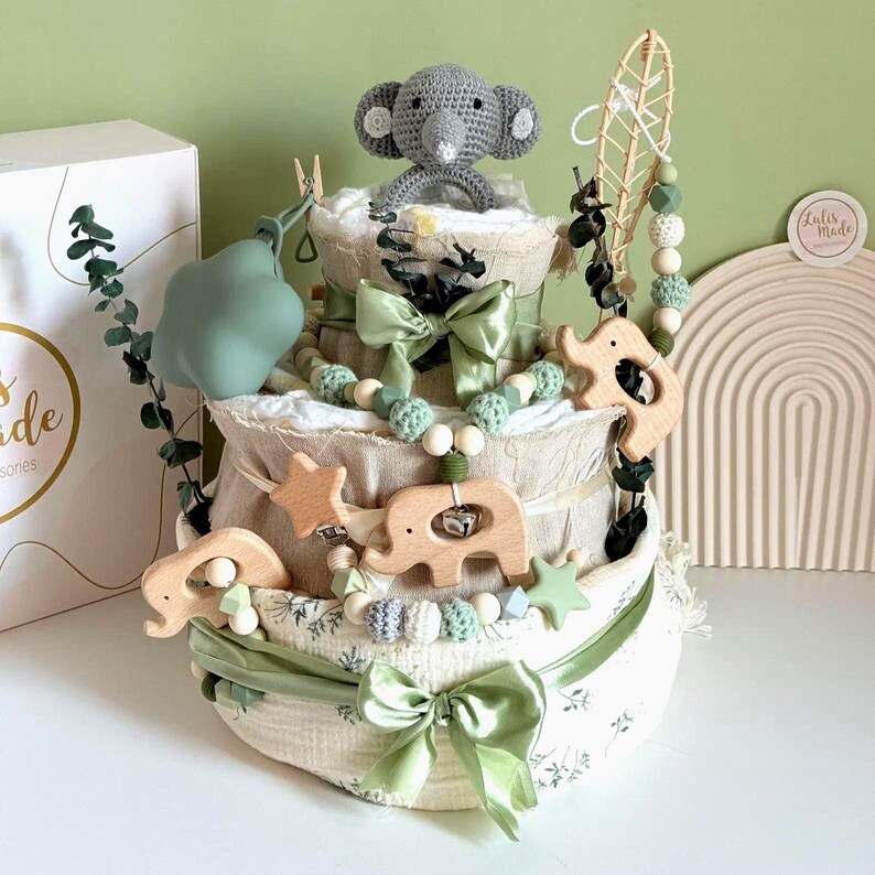 May include: A three-tiered baby diaper cake decorated with green bows, wooden elephant and star accents, and a crocheted gray elephant topper. The cake is adorned with a garland of wooden beads and green accents.