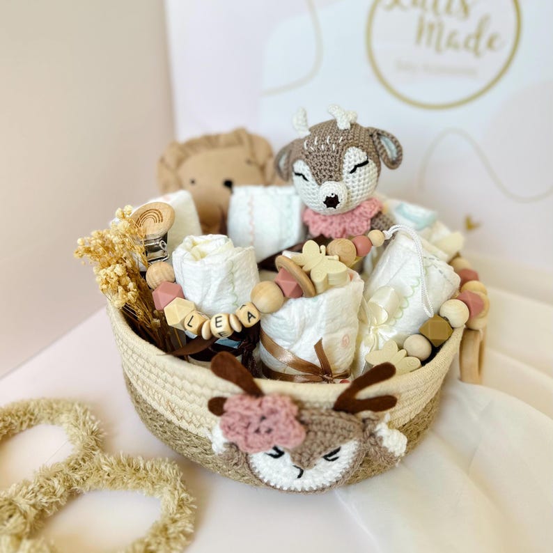 May include: A wicker basket filled with baby items, including a crocheted deer toy, a wooden teether, and a set of white washcloths. The basket is decorated with a crocheted deer head and a pink flower.