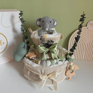 May include: A two-tiered diaper cake with a gray crocheted elephant topper. The cake is decorated with green ribbons, wooden beads, and a wooden star and elephant. Eucalyptus sprigs and a green pear-shaped object add to the decor.