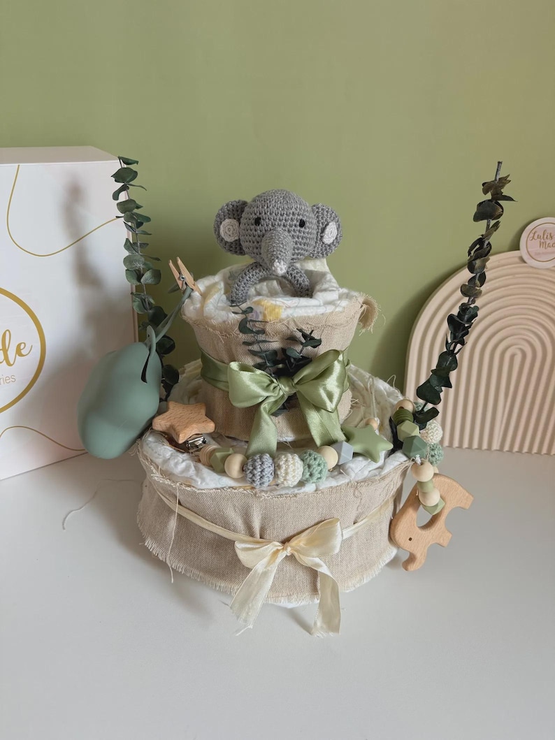 May include: A two-tiered diaper cake with a gray crocheted elephant topper. The cake is decorated with green ribbons, wooden beads, and a wooden star and elephant. Eucalyptus sprigs and a green pear-shaped object add to the decor.