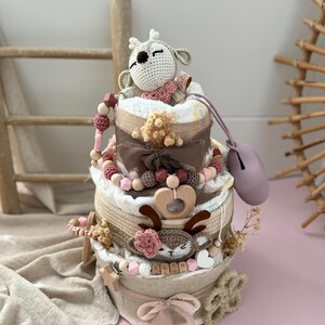 May include: A three-tiered diaper cake decorated with a crocheted deer toy, wooden beads, and a pink ribbon. The cake is wrapped in burlap and has a wooden heart-shaped ring on the middle tier. The bottom tier has a wooden name tag with the name "ELLIN" on it.