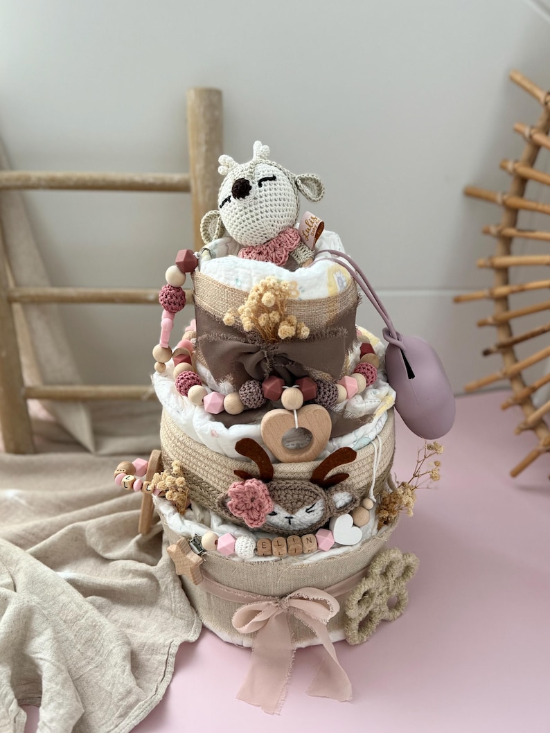 May include: A three-tiered diaper cake decorated with a crocheted deer toy, wooden beads, and a pink ribbon. The cake is wrapped in burlap and has a wooden heart-shaped ring on the middle tier. The bottom tier has a wooden name tag with the name "ELLIN" on it.