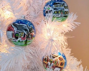 May include: Three hand-painted Christmas ornaments featuring houses with snow-covered roofs and a blue, green, and yellow color scheme. The ornaments are hanging on a white artificial Christmas tree.