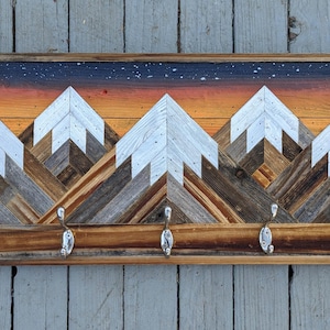 May include: Wooden wall-mounted coat rack featuring a mountain scene. The artwork depicts white mountain peaks against a sunset sky with a starry night. Includes four silver-toned hooks for hanging items. The frame is made of natural wood.