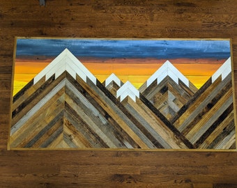 May include: A wooden wall art piece depicting a mountain range with a sunset sky. The mountains are made of various shades of wood and painted white at the peaks. The sky is painted in shades of blue, orange, and yellow.
