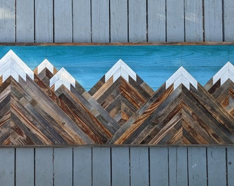 May include: A wooden wall art piece depicting a mountain range with a turquoise blue sky. The mountains are made of reclaimed wood and have white peaks.
