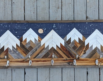 May include: A wooden wall-mounted coat rack with a mountain range design. The mountains are made of reclaimed wood and painted white. The background is painted dark blue with white speckles to resemble a starry night sky. The rack has five metal hooks.