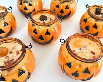 Puede incluir: Varios candelabros de vidrio naranja con forma de calabazas de Halloween con caras negras. Cada candelabro tiene un asa de metal negro y está lleno de cera de color claro y piedras decorativas. Las velas están dispuestas sobre una superficie blanca.