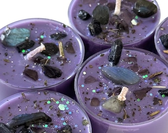 May include: Close-up of several small, round, purple candles. Each candle is decorated with small crystals, dark stones, and glitter. The wicks are white and appear to be made of cotton. The candles are arranged closely together.