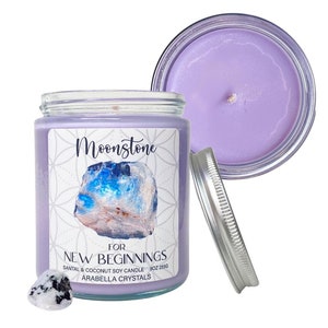 May include: A lavender soy candle in a glass jar with a silver lid. The label says "Moonstone for New Beginnings Santal & Coconut Soy Candle 10oz Arabella Crystals". The candle is topped with a small moonstone crystal.