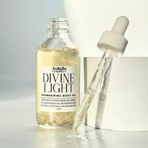 Könnte beinhalten: Eine durchsichtige Glasflasche mit einer goldenen, schimmernden Flüssigkeit. Die Flasche ist mit "Arabella + Crystals + Divine Light Shimmering Body Oil" und "Made with sunflower oil, jojoba oil, grapeseed oil, biodegradable glitter, crystals, and fragrance 2 oz" beschriftet.
