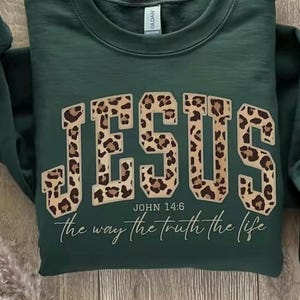 May include: A dark green sweatshirt featuring the word "JESUS" in large leopard print lettering. The text "JOHN 14:6" and the phrase "the way the truth the life" are below the word "JESUS" in a cursive font.