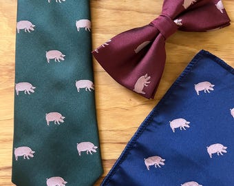 Könnte beinhalten: Ein Set Herrenaccessoires mit einer grünen Krawatte, einer weinroten Fliege und einem marineblauen Einstecktuch. Jedes Teil ist mit hellrosa Schweine-Illustrationen versehen. Die Artikel sind auf einer Holzoberfläche angeordnet.