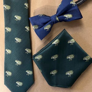 Puede incluir: Un conjunto de corbata verde oscuro, pajarita azul marino y pañuelo de bolsillo verde oscuro. Cada artículo presenta un patrón repetido de ranas verdes. La pajarita tiene un tejido de espiga. La corbata es de ancho estándar.