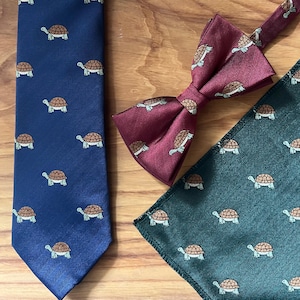 Peut inclure: Une cravate bleu marine, un nœud papillon bordeaux et une pochette verte, tous avec un motif de tortue. La cravate a un tissage en chevrons. Le nœud papillon et la pochette sont bordeaux avec un motif de tortue.