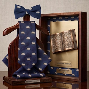 May include: A dark blue bow tie, necktie, and pocket square set with a repeating light-colored open book pattern displayed on a wooden hanger stand. Beside it, a framed bronze-colored open book relief with plaques reading OPEN BOOK PORTRAIT.