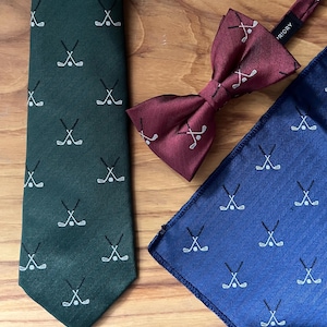May include: A green tie, burgundy bow tie, and blue pocket square with a golf club and ball pattern. The bow tie has "PRIORY" printed on the band. The items are arranged on a wooden surface.