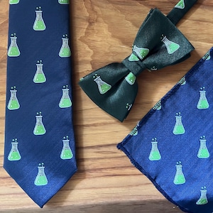 May include: A navy blue tie, bow tie, and pocket square set. Each item features a repeating pattern of green lab flasks. The bow tie is dark green. The pocket square has a blue herringbone pattern.