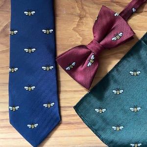 May include: A navy blue tie, burgundy bow tie, and green pocket square, each featuring a repeating bee motif. The tie has a herringbone weave. The bow tie is pre-tied. The pocket square has a similar weave to the tie.
