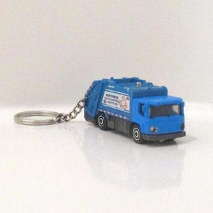 May include: A blue toy garbage truck keychain. The truck has black wheels and a silver chain and keyring. The text on the side of the truck reads "Matchbox Valley Recycling & Recycling".