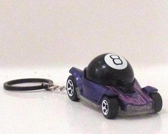May include: A miniature purple toy car with a black 8-ball on top. The car has black wheels and a silver keychain ring attached. The 8-ball has a white number 8 on it. The car is a novelty item.
