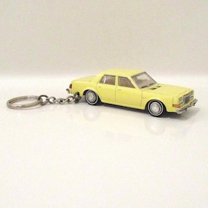 May include: A yellow toy car keychain. The miniature car has a classic sedan design with chrome accents and white-walled tires. The keychain ring and chain are silver-colored.