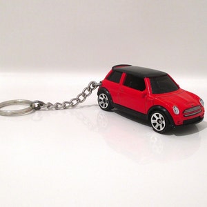 May include: Red and black miniature car keychain. The toy car has a black roof, red body, and silver chain with a keyring. The car has black tires with silver rims. The keychain is on a white background.