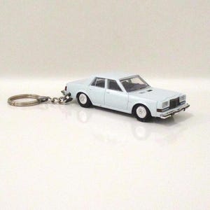 May include: A light blue toy car keychain. The miniature car has four doors, white wheels, and a silver-colored front grill. The keychain ring and chain are also silver. The car is a classic sedan style.