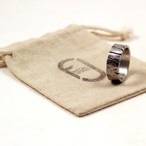 Silver pewter ring 8mm wide with deep cuts and hammered texture sitting on edge on recycled cotton gift bag stamped with logo