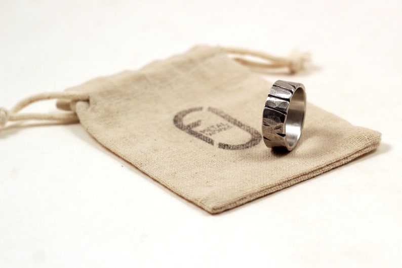 Silver pewter ring 8mm wide with deep cuts and hammered texture sitting on edge on recycled cotton gift bag stamped with logo