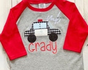 May include: A gray and red long-sleeved shirt with a black and white checkered police car applique. The car has a blue light on top and the name "Grady" is embroidered below it.