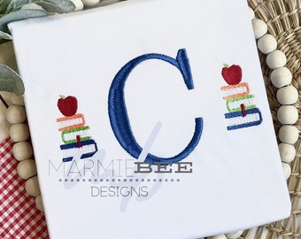 May include: A white t-shirt with a blue embroidered letter C and two stacks of books with red apples on top. The text "MARMIE BEE DESIGNS" is embroidered below the letter.