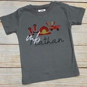 May include: A gray t-shirt with a design featuring a firefighter's helmet, fire truck, and fire hoses. The name "Nathan" is written in cursive below the design. The shirt is made of a soft material.