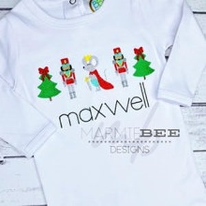 May include: White long-sleeve onesie with a Christmas theme featuring a Nutcracker, a mouse king, and Christmas trees. The onesie is personalized with the name "maxwell".
