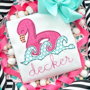 Op de afbeelding: Wit T-shirt met een roze flamingo-ontwerp dat zwemt in blauwe en witte stippelgolven. De naam "decker" is geborduurd in roze script. Een turkooizen strik en roze en zwart gestreept lint zijn op de achtergrond.