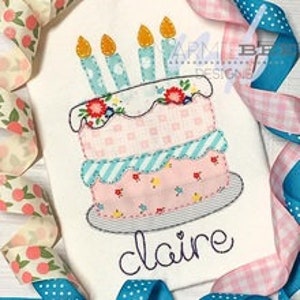 Könnte beinhalten: Ein gestickter Entwurf eines rosa und blauen Geburtstagskuchens mit Kerzen und Blumen auf einem weißen Stoff. Der Name "claire" ist unter dem Kuchen gestickt.