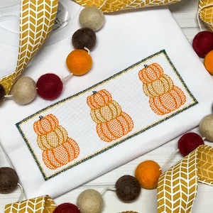 May include: White baby onesie with a green and gold embroidered design of three stacked pumpkins. The pumpkins are orange, yellow, and brown.
