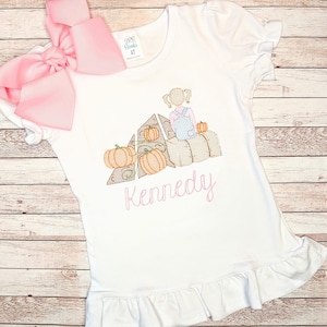 Puede incluir: Camiseta blanca con un dobladillo de volantes rosa con un diseño bordado de una niña sentada en pacas de heno con calabazas. El texto "Kennedy" está bordado debajo del diseño.