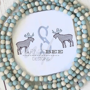 Op de afbeelding: Witte stof met twee geborduurde elanden en de tekst "MARMIE BEE DESIGNS". De stof is omgeven door een touw van blauwe en witte kralen.