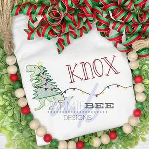 May include: White t-shirt with a green dinosaur holding a Christmas tree with lights. The dinosaur has a star on its head. The name "Knox" is embroidered in red above the dinosaur and a string of lights is embroidered below. The text "Parmie Bee Designs" is embroidered below the lights.