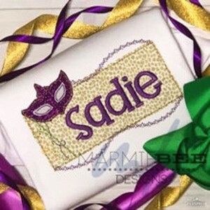 May include: A white t-shirt with a purple and gold Mardi Gras themed design. The design features a purple mask and the name "Sadie" in purple text.