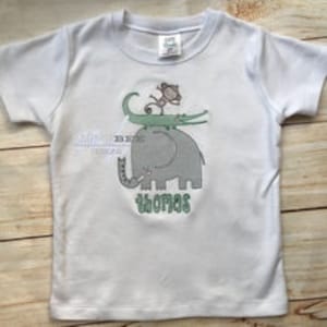 Peut inclure: T-shirt à manches courtes blanche avec un motif vert, marron et gris d'un singe chevauchant un crocodile chevauchant un éléphant. Le texte "Whoa!" est imprimé sous les animaux.
