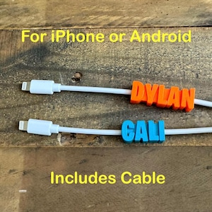May include: Two white charging cables with orange and blue personalized name tags that say "DYLAN" and "CALI". The text "For iPhone or Android" and "Includes Cable" is written in yellow on a wooden background.