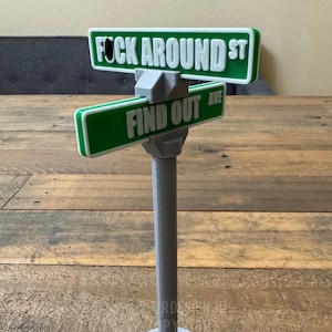 May include: A gray street sign post with two green street signs. The top sign reads "F**K AROUND ST" and the bottom sign reads "FIND OUT AVE".