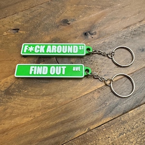 May include: Two green street sign keychains with white text. The top keychain says "F*CK AROUND ST" and the bottom keychain says "FIND OUT AVE".