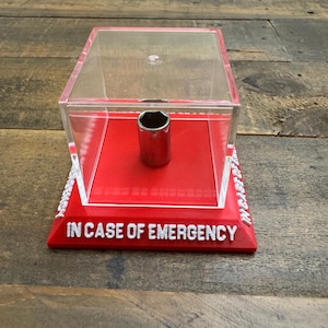 May include: A chrome socket wrench is displayed inside a clear acrylic box with a red base. The base features the white text "IN CASE OF EMERGENCY". The box is placed on a wooden surface.