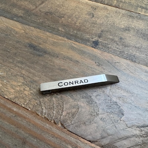 May include: A rectangular, silver-colored metal object with the name "CONRAD" printed in black. The object has a beveled edge on one end. The item is resting on a wooden surface.