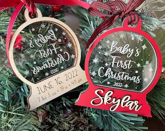 May include: Two wooden Christmas ornaments with clear glass inserts. The ornament on the left is natural wood with the text "Baby's First Christmas" and the date "June 16, 2022" and birth stats. The ornament on the right is red with the text "Baby's First Christmas 2022" and the name "Skylar".