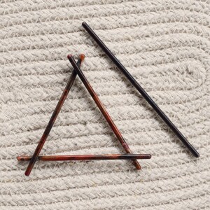 May include: Three hair sticks arranged in a triangle shape on a white textured surface. Two of the sticks are brown with red and black accents, and the third stick is black.