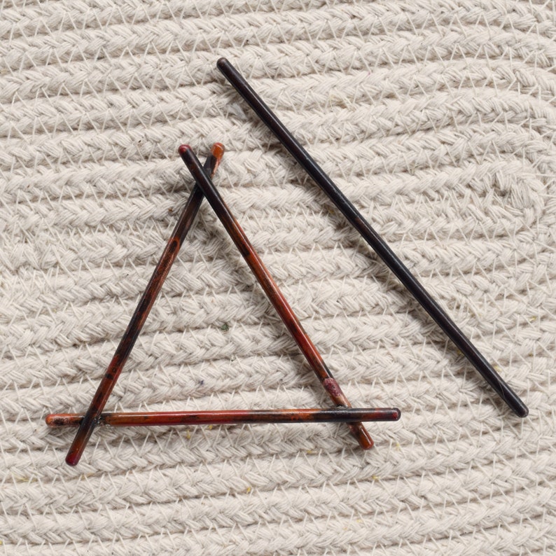 May include: Three hair sticks arranged in a triangle shape on a white textured surface. Two of the sticks are brown with red and black accents, and the third stick is black.